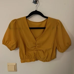 Gul croppad blus med puffärm - Trendig gul blus med korta puffärmar och v-ringning. Blusen är croppad med rynkade detaljer framtill och resår i nederkant. Perfekt för dig som vill ha en färgklick i garderoben.
