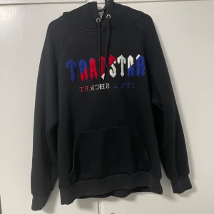 Trapstar hoodie  - Säljer en svart hoodie från Trapstar med broderad text i blått, rött och vitt på bröstet. Tröjan har huva  och en stor magficka. Perfekt för dig som gillar streetwear och vill sticka ut lite extra.