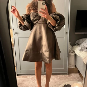 Brun axelbandslös klänning med puffärmar - Säljer en brun axelbandslös klänning med markerad byst och stora puffärmar. Klänningen har en lätt glansig finish och är sydd i ett satinliknande material. Endast använd så i toppskick! Nypris: 3000kr