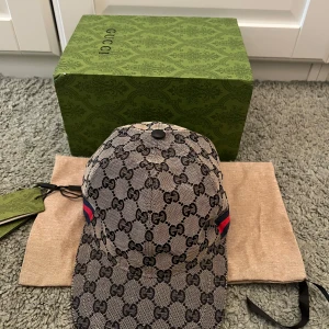 Gucci keps med monogram och röd-blå detalj - Säljer en exklusiv Gucci keps i beige med svart monogrammönster och röd-blå rand på sidan. Kepsen är i storlek L (59 cm) och har justerbart spänne bak. Perfekt accessoar för dig som gillar lyxiga märken.