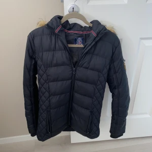 Svart quiltad jacka med huva från Vinson Polo Club - Snygg svart quiltad jacka från Vinson Polo Club med huva och fuskpälsdetalj. Jackan har dragkedja framtill, två fickor och en liten ficka på ärmen med dragkedja och sportig känsla. Perfekt för lite kyliga dagar och enkel att matcha med olika outfits.