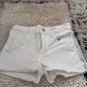 Vita jeansshorts med paljettdetalj från H&M - Snygga vita jeansshorts med råa kanter och en glittrig paljettremsa längs sidorna. Klassisk femficksmodell med knapp och dragkedja framtill. Perfekta för sommarens alla äventyr!