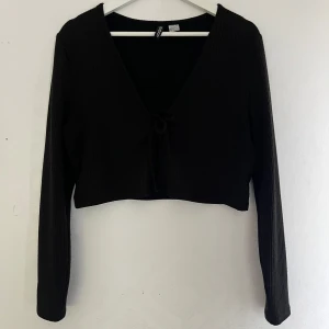 Svart croppad ribbad kofta med knyt - Säljer en svart croppad kofta med lång ärm och djup v-ringning. Koftan är ribbad och har knyt framtill, den är väl använd så den är lite nopprig. 