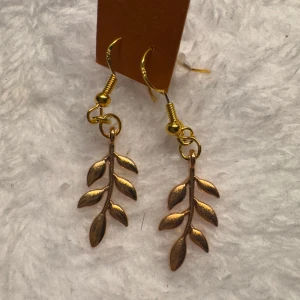 Guldiga örhängen med bladmotiv - Säljer ett par snygga örhängen i guld med hängande bladformade detaljer. Perfekta för dig som gillar naturinspirerade accessoarer och vill ha något unikt till din stil. Krokarna är i sterling silver