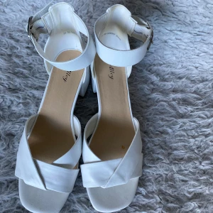 Vita sandaler med klack och spänne - Säljer ett par vita sandaler med bred klack och öppen tå. Skorna har en snygg korsad rem framtill och justerbart spänne vid ankeln. Perfekta för vår och sommar!