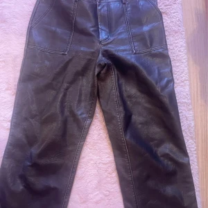 Svarta skinnbyxor från Zara - Snygga svarta skinnbyxor från Zara med raka ben och stora fickor framtill. Byxorna har hög midja och är tillverkade i ett glansigt skinnmaterial som ger en cool look. Perfekta att styla med en enkel topp eller stickad tröja.