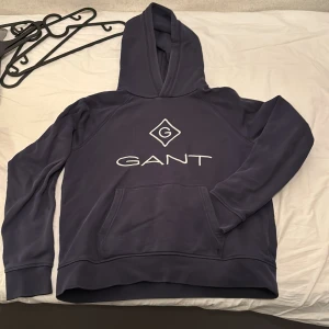 Mörkblå hoodie från GANT - Säljer en mörkblå hoodie från GANT med stor vit logga framtill. Tröjan har huva och en stor magficka. Perfekt för dig som gillar klassisk och stilren design.