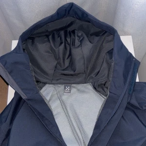 Mörkblå vindjacka med huva från Haglöfs - Säljer en mörkblå vindjacka från Haglöfs med huva och dragkedja. Jackan har meshfoder på insidan och en stilren design. Perfekt för blåsiga dagar och enkel att matcha med olika outfits.