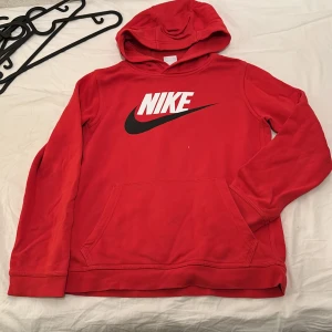 Röd hoodie från Nike - Säljer en röd hoodie från Nike med klassisk logga i vitt och svart på bröstet. Tröjan har huva och en stor magficka framtill. Perfekt för en sportig och avslappnad stil.