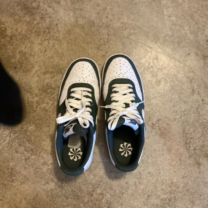 Nike Air Force 1 i vitt och grönt - Säljer ett par Nikeskor med vit bas och gröna detaljer. Skorna har klassisk rund tå, perforerad tåbox och vit sula. Swoosh-logga på sidan och vita skosnören. Perfekta för dig som gillar stilrena sneakers med en twist.