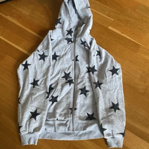 Grå hoodie med svarta stjärnor från Lindex - Säljer en grå hoodie från Lindex med svarta stjärnor över hela tröjan. Tröjan har dragkedja framtill, huva och två fickor. Perfekt för dig som gillar en cool och avslappnad stil.