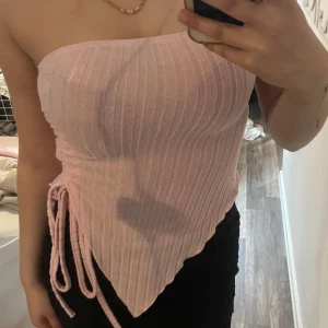 Ljusrosa ribbad tubtopp med knyt - Säljer en ljusrosa ribbad tubtopp med knyt i sidan och asymmetrisk nederkant. Toppen är axelbandslös och har en snygg croppad passform som framhäver figuren. Perfekt för varma dagar eller när du vill sticka ut lite extra.