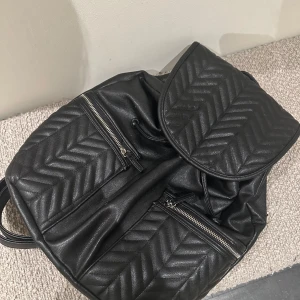 Svart Don Donna ryggsäck med quiltad design - Säljer en svart ryggsäck från Don Donna i skinnimitation med quiltad mönster på locket och framsidan. Väskan har två dragkedjefickor framtill. Perfekt för dig som vill ha en stilren och praktisk accessoar.