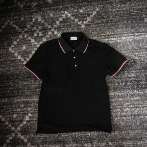 Svart pikétröja från Moncler Medium Polo - Säljer en svart pikétröja från Moncler med klassisk krage och knäppning framtill. Kragen och ärmsluten har snygga ränder i vitt och rött för en sportig look. Perfekt för dig som gillar stilrena och exklusiva plagg.