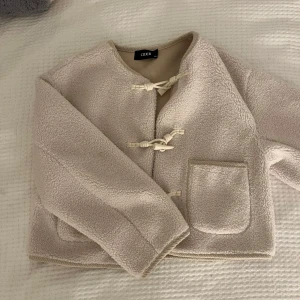 Beige teddyjacka från Cider - Mysig beige teddyjacka från Cider med två stora fickor framtill och dekorativa knytknappar. Jackan har lång ärm och är tillverkad i ett mjukt, fleece-liknande material. Perfekt för lager-på-lager och en avslappnad stil.