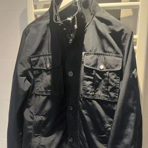 J.Lindeberg Field Jacket Navy - Stilren Field Jacket från J.Lindeberg i mörkblått, storlek M, i mycket fint skick. En mångsidig jacka med både funktion och design – passar utmärkt till både jeans och kostymbyxor. Perfekt jacka för övergångsperioder.