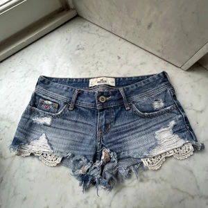 Blå jeansshorts med spets från Hollister - Säljer ett par blå jeansshorts från Hollister med slitna detaljer och fransar. Shortsen har spetskant nedtill och klassisk femficksdesign. Perfekta för sommaren och har låg midja. 
