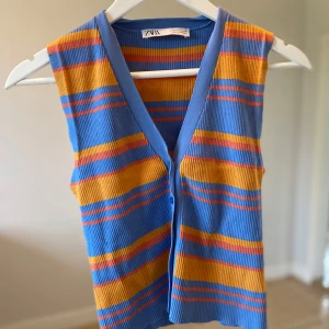 Randig stickad väst från Zara - Säljer en färgglad stickad väst från Zara med blå, orange och röda ränder. Västen har v-ringning och knappar framtill. Perfekt att styla över en t-shirt eller skjorta för en trendig look.