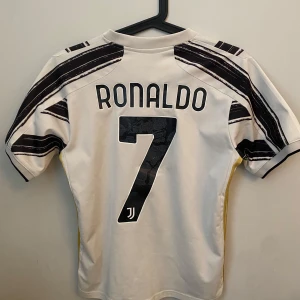 Juventus fotbollströja med Ronaldo där bak  - Juventus hemmatröja 2020/21 med Cristiano Ronaldo på ryggen. Vit bas med svarta och guldiga ränder. Adidas-märke på bröstet. Perfekt för fans av både klubben och spelaren.