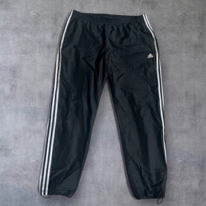 Svarta träningsbyxor från Adidas med vita ränder - Svarta träningsbyxor från Adidas med klassiska vita ränder längs sidorna och liten Adidas-logga på låret. Byxorna har resår i midjan och dragsko samt resår vid bensluten. Perfekta för sport eller chill. Matchande jackan är i min profil