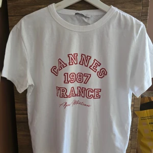 Vit t-shirt med tryck från Zara - Vit t-shirt från Zara med rött tryck på bröstet där det står 'Cannes 1987 France'. Klassisk rund halsringning och korta ärmar. Perfekt för en avslappnad stil.
