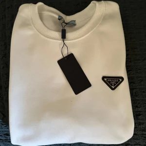 Vit sweatshirt från Prada - Säljer en stilren vit sweatshirt från Prada med klassisk rund halsringning och diskret logotyp på bröstet. För en skön sommar look! Helt ny! Inte använd! Storlek S, för mer bilder / info hör av er 🤗
