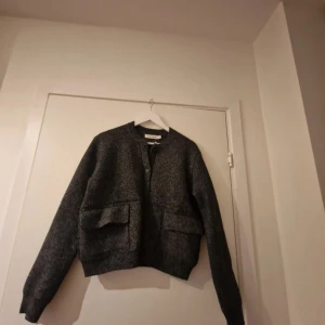Mörkgrå cardigan jacka med fickor - Säljer en mörkgrå cardigan jacka med två stora fickor framtill och ribbade muddar. Jackan har en enkel, stilren design utan synliga knappar eller dragkedja. Perfekt för lager-på-lager och en avslappnad look.