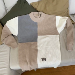 Beige och grå stickad tröja från Burberry - Säljer en stickad tröja från Burberry i blockmönster med beige, grå och vita partier. Tröjan har rund halsringning, långa ärmar och en liten broderad Burberry-logga nedtill. Perfekt för dig som gillar stilrena färger och klassisk design.