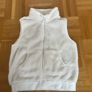 Vit pälsväst från H&M - Mysig vit väst i mjuk fuskpäls från H&M. Västen har dragkedja framtill, två fickor och hög krage. Perfekt för lager-på-lager och ger en fluffig känsla.