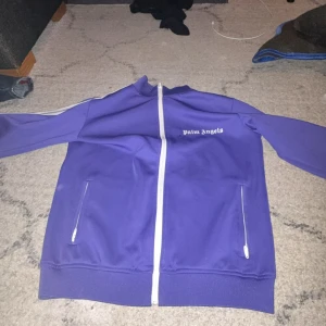 Lila track jacket från Palm Angels - Säljer en lila track jacket från Palm Angels med vit dragkedja och vita detaljer på ärmarna. Jackan har ståkrage och två fickor med dragkedja framtill. Perfekt för en sportig och trendig look.