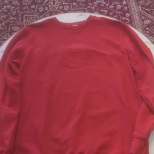 Röd sweatshirt från Moncler - Säljer en röd sweatshirt från Moncler med ton-i-ton broderad logga på bröstet och en matchande logotyp-patch på ärmen. Tröjan har rund halsringning och långa ärmar. Perfekt för dig som gillar stilrena märkesplagg.
