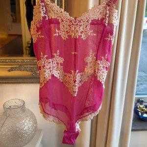 Intimissimi body  - Säljer en snygg rosa body med tunna axelband och vackra blommiga spetsdetaljer i beige. Bodyn är genomskinlig och har en feminin, romantisk look med spets längs kanterna. 
