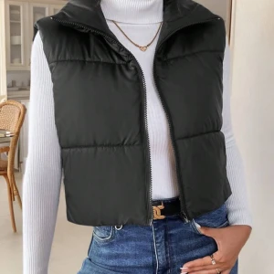 Svart dunväst med dragkedja - Snygg svart dunväst med hög krage och dragkedja framtill. Västen är croppad och har en stilren, enkel design som passar perfekt över en tröja eller hoodie. 