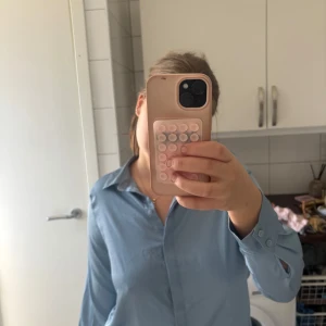 Ljusblå skjorta från H&M - Säljer en klassisk ljusblå skjorta från H&M. Skjortan har lång ärm, krage och knäppning framtill. Perfekt att styla till jeans eller kjol för en clean look. Materialet känns mjukt och lätt mot huden.