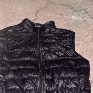 Svart dunväst från Emporio Armani EA7 - Snygg svart dunväst från Emporio Armani EA7 med quiltad design och dragkedja framtill. Västen har hög krage och diskret logga på bröstet. Perfekt för lager-på-lager och stilrena outfits.