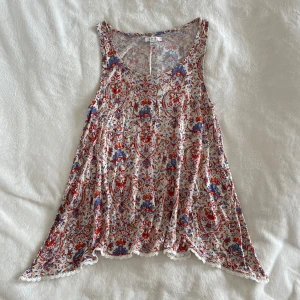 Mönstrad ärmlös linnetopp från Amisu - Säljer en luftig och flowy ärmlös linnetopp från Amisu med ett färgglatt blommigt och paisley-inspirerat mönster i rött, blått och beige på vit botten. Toppen har spetsdetaljer längs nederkanten och rund halsringning.