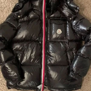 Moncler Montbelliard - Säljer en svart glansig dunjacka från Moncler med avtagbar huva och klassisk logga på bröstet. Jackan har dragkedja med röd, vit och blå detalj samt flera fickor framtill. NFC SCAN och qr kod samt tags medföljer. 