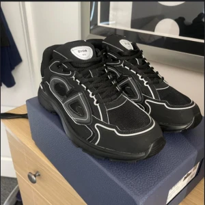 Dior B30 - Säljer ett par svarta Dior B30 sneakers med vita detaljer och reflekterande inslag. Skorna har snörning, rund tå och en chunky platt sula. Perfekta för dig som gillar streetwear och vill sticka ut med en lyxig touch.