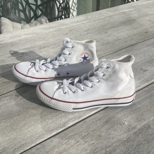 Vita Converse All Star high-top sneakers - Klassiska vita Converse All Star high-top sneakers med röd och blå rand längs sulan. Skorna har snörning framtill, rund tå och är tillverkade i canvas. Perfekta för en avslappnad och stilren look. Dem är nästan oanvända! 