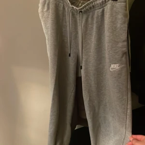 Grå mjukisbyxor från Nike - Säljer ett par grå mjukisbyxor från Nike med dragsko i midjan och logga på vänster ben. Byxorna har en avslappnad passform och är perfekta för chill eller träning. Klassisk design med resår i midjan och benslut.