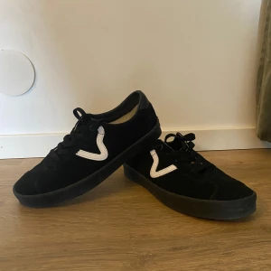 Svarta sneakers från Vans med vit detalj - Säljer ett par svarta Vans sneakers i mocka med vit V-formad detalj på sidan. Skorna har snörning och en klassisk gummisula med våffelmönster undertill. Perfekta för dig som gillar stilrena och tidlösa sneakers.