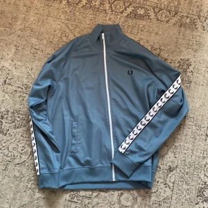 Blå track jacket från Fred Perry - Säljer en blå track jacket från Fred Perry med vita detaljer och logga på bröstet. Jackan har vita band med Fred Perry-loggor längs ärmarna och dragkedja framtill. Använd men inga defekter alls. 