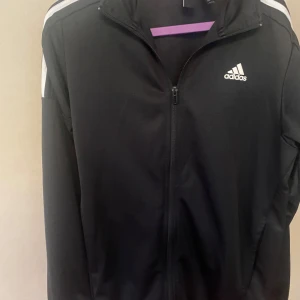 Svart träningsjacka från Adidas - Svart träningsjacka från Adidas med vit logga på bröstet och vita ränder på axlarna. Jackan har dragkedja framtill och hög krage. Perfekt för sportiga tillfällen och enkel att matcha med andra plagg.