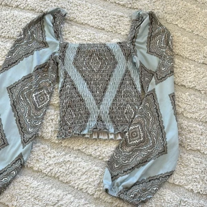 Mönstrad blus med puffärmar - Säljer en ljusblå blus med brunt och vitt paisleymönster. Toppen har smockad kropp, fyrkantig ringning och långa puffärmar med resår vid handlederna. Perfekt för dig som gillar bohemisk stil!
