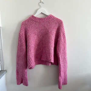 Rosa stickad croppad tröja - Säljer en rosa stickad tröja med croppad passform och långa ärmar. Tröjan har en bred ribbad struktur och hög halsringning. Perfekt för att addera färg till din garderob!