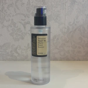 COSRX Advanced Snail 96 Mucin Power Essence - En genomskinlig pumpflaska med COSRX Advanced Snail 96 Mucin Power Essence. Innehåller 100 ml essence med snigelsekret, perfekt för återfuktning och glow. Flaskan är frostad och har svart etikett med beige detaljer.
