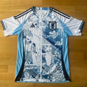 Japan fotbollströja Adidas x Dragon Ball - Unik Japan fotbollströja från Adidas med Dragon Ball-motiv i blått och vitt 2024/2025. Tröjan har korta ärmar, svart JFA-märke på bröstet och coola manga-inspirerade tryck över hela plagget. Perfekt för dig som gillar en speciell fotbollströja inför sommaren 