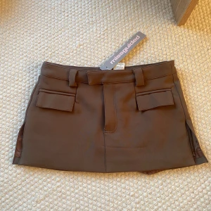 Brun minikjol med fickor från Missguided - Brun kjol med inbyggda shorts. Fickor fram. Storlek S! Aldrig använd med lappar kvar. Köpt för 400kr