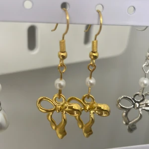 Guldiga örhängen med rosett och pärla - Snygga örhängen i guld med dekorativa rosetter och en liten vit pärla ovanför. Perfekta för att ge din outfit en söt och trendig touch.