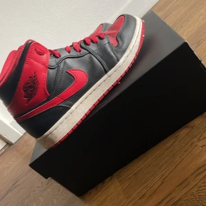 Nike Air Jordan 1 mid Svart/Röd - Säljer ett par klassiska Nike Air Jordan 1 High i svart och rött med vita sulor. Skorna har högt skaft, snörning och den ikoniska swoosh-loggan på sidan. Perfekta för dig som gillar streetwear och sneakers med retrostil.